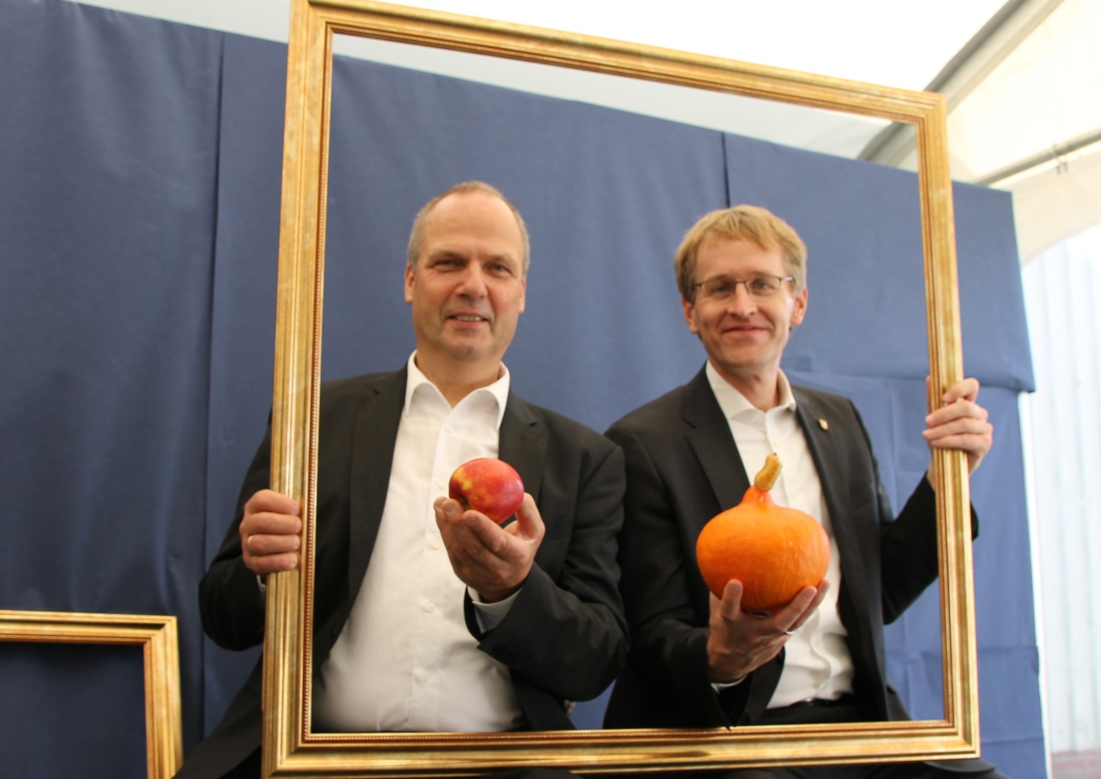 Werner Schwarz, Landwirtschaftsminister von Schleswig-Holstein, und der Ministerpräsident des Landes, Daniel Günther, halten einen Bilderrahmen und Kürbisse in ihren Händen.